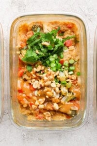 A glass container with a dish featuring peanut chicken and vegetables, topped with chopped peanuts, green onions, and cilantro, set on a textured surface.