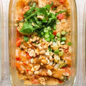 A glass container with a dish featuring peanut chicken and vegetables, topped with chopped peanuts, green onions, and cilantro, set on a textured surface.