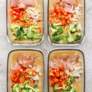 Four glass meal prep containers filled with uncooked peanut chicken, diced peppers, onions, broccoli, and sauce rest on a textured surface.