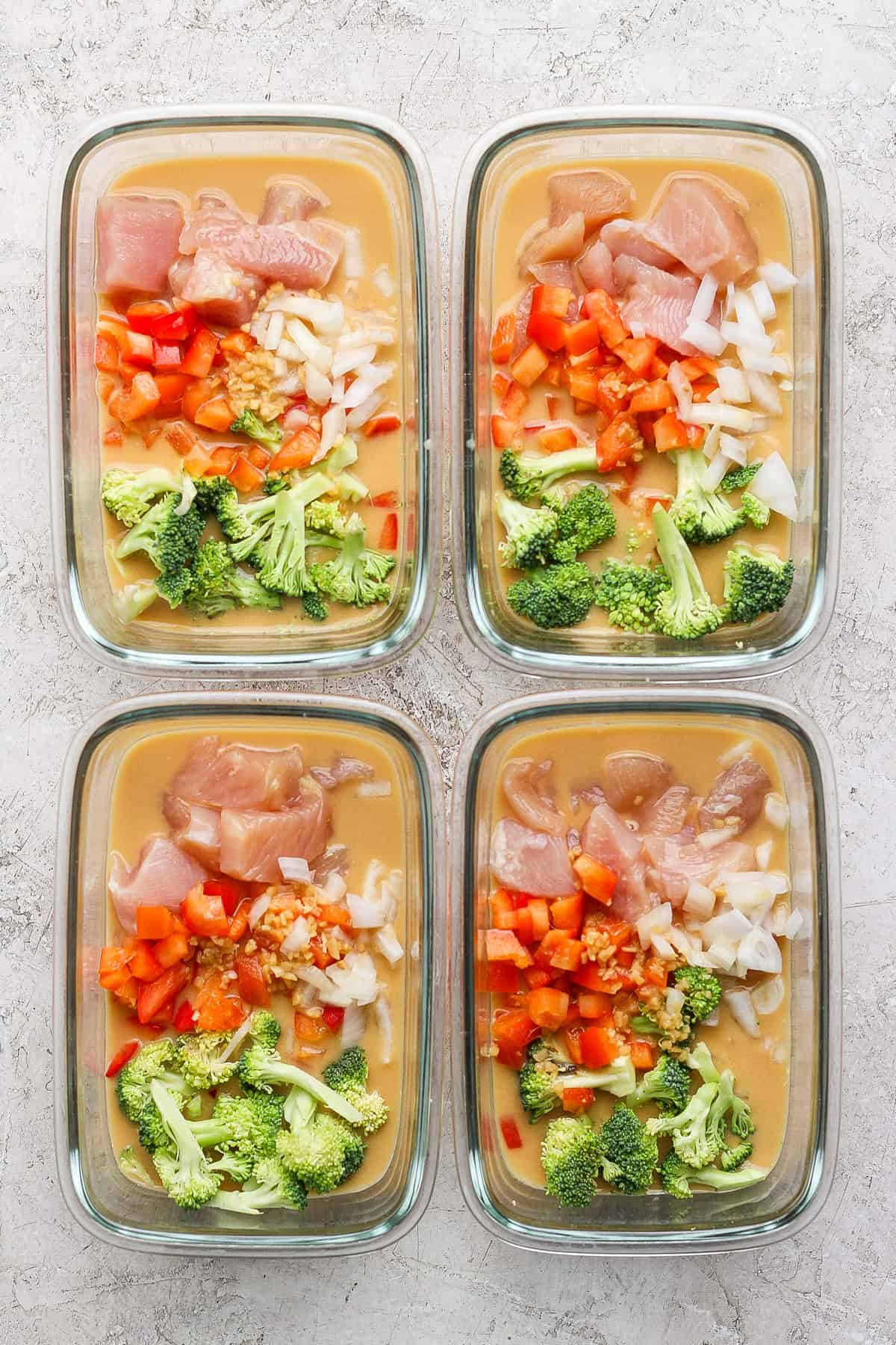 Four glass meal prep containers filled with uncooked peanut chicken, diced peppers, onions, broccoli, and sauce rest on a textured surface.