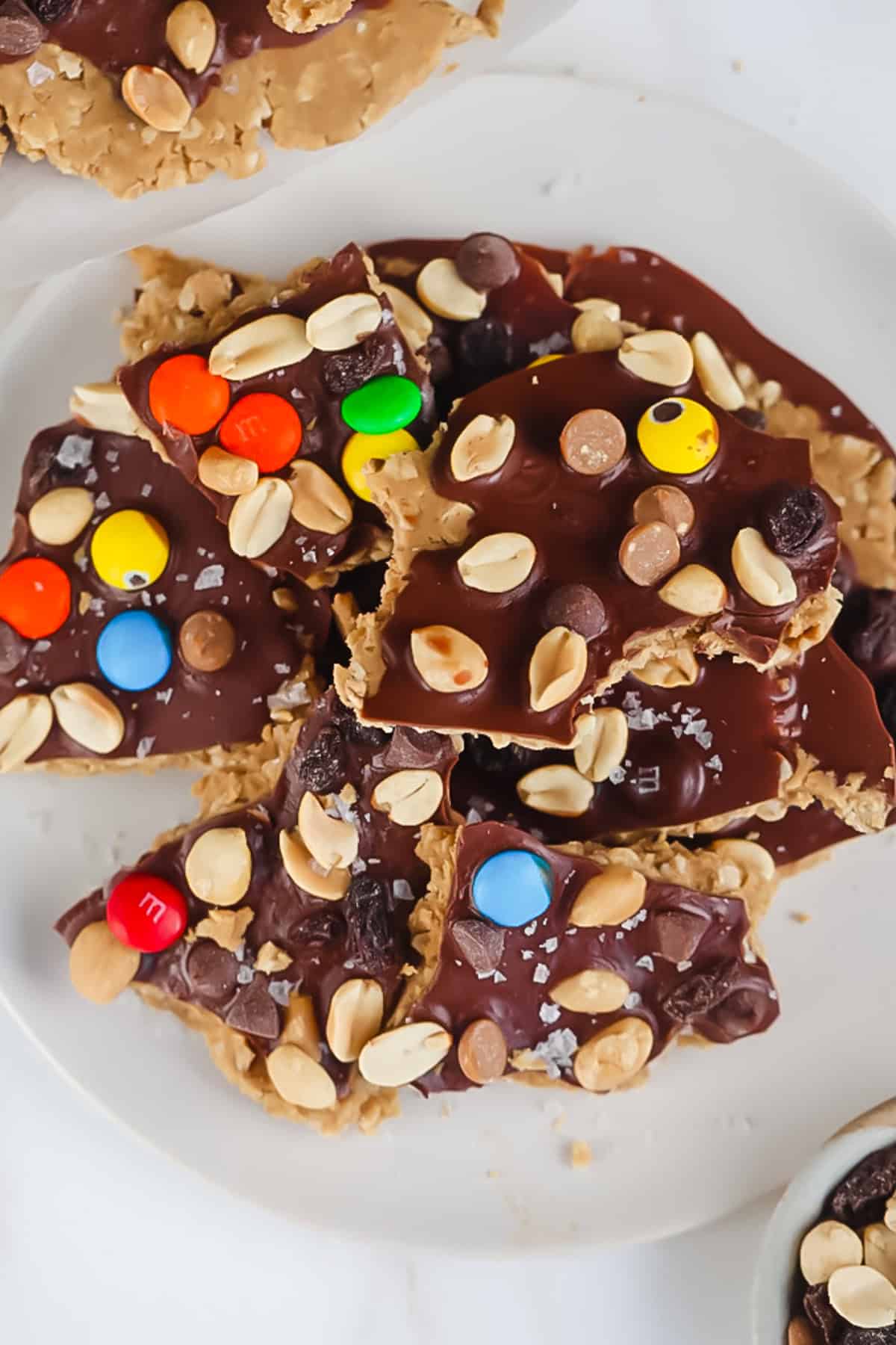 A plate of peanut butter and chocolate bark topped with colorful candy-coated chocolates, peanuts, and sea salt flakes, on a white surface.