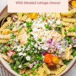 A bowl of pasta salad with fusilli, peas, red onions, cherry tomatoes, and a creamy dressing, topped with blended cottage cheese and herbs.