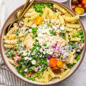 A bowl of pasta salad with fusilli, cherry tomatoes, peas, feta cheese, and red onion. Two gold utensils rest on the side. Small bowls of red onions and cherry tomatoes are nearby.