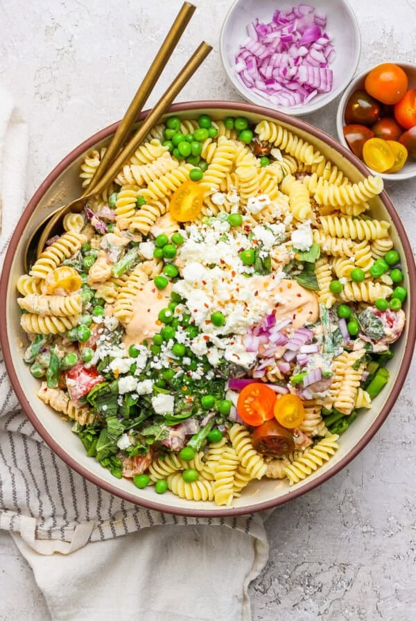 A bowl of pasta salad with fusilli, cherry tomatoes, peas, feta cheese, and red onion. Two gold utensils rest on the side. Small bowls of red onions and cherry tomatoes are nearby.