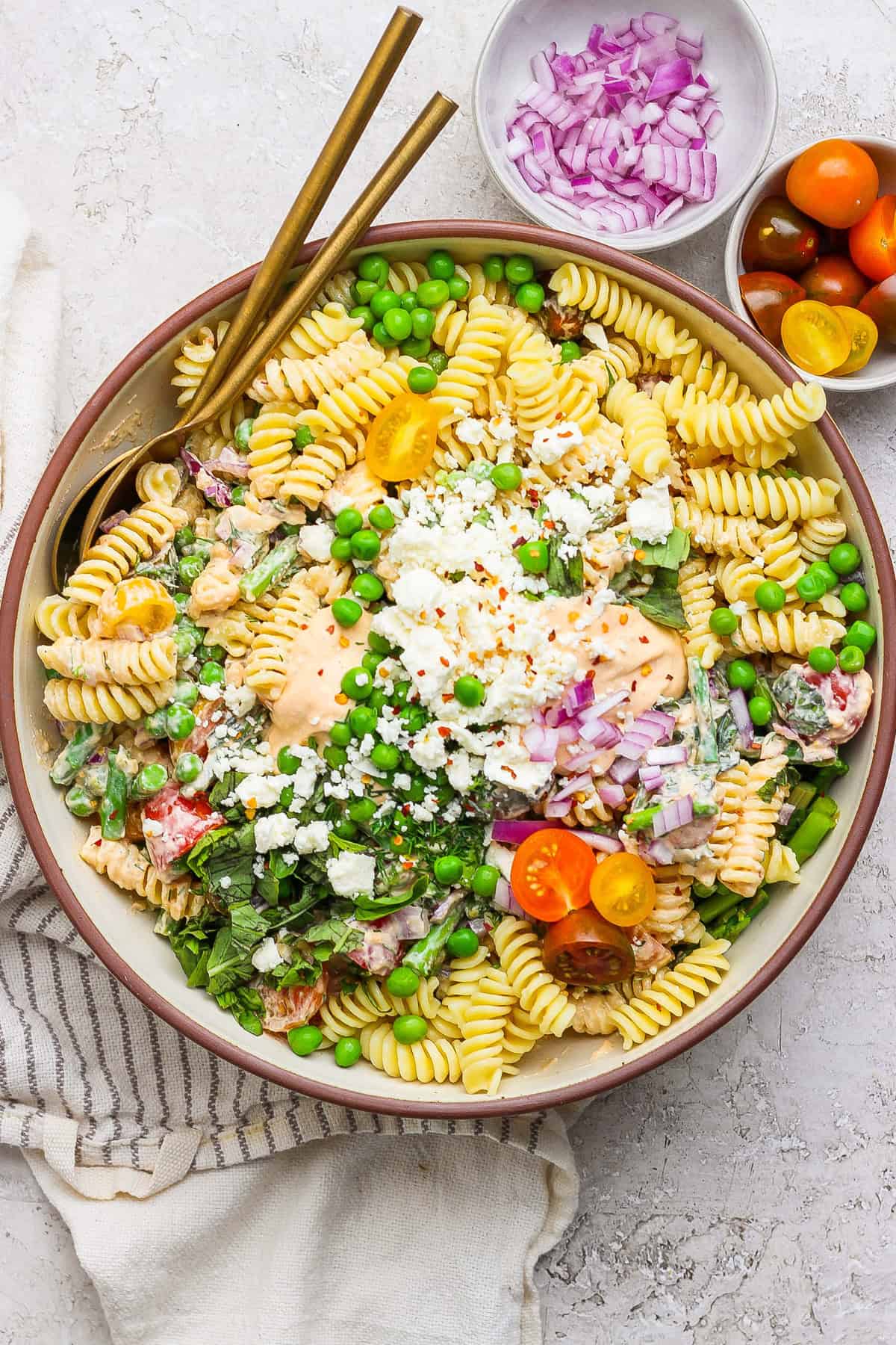 A bowl of pasta salad with fusilli, cherry tomatoes, peas, feta cheese, and red onion. Two gold utensils rest on the side. Small bowls of red onions and cherry tomatoes are nearby.