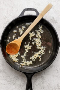 A cast iron skillet with sautéing minced garlic in oil, accompanied by a wooden spoon.