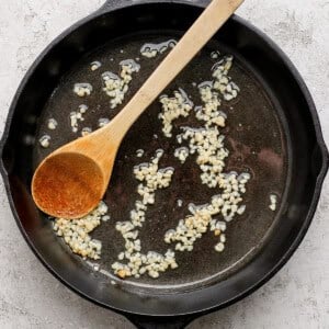 A cast iron skillet with sautéing minced garlic in oil, accompanied by a wooden spoon.