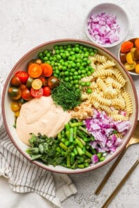 A bowl with rotini pasta, cherry tomatoes, peas, chopped red onion, green beans, fresh herbs, and a creamy sauce. A small bowl of onions and cutlery are beside it.