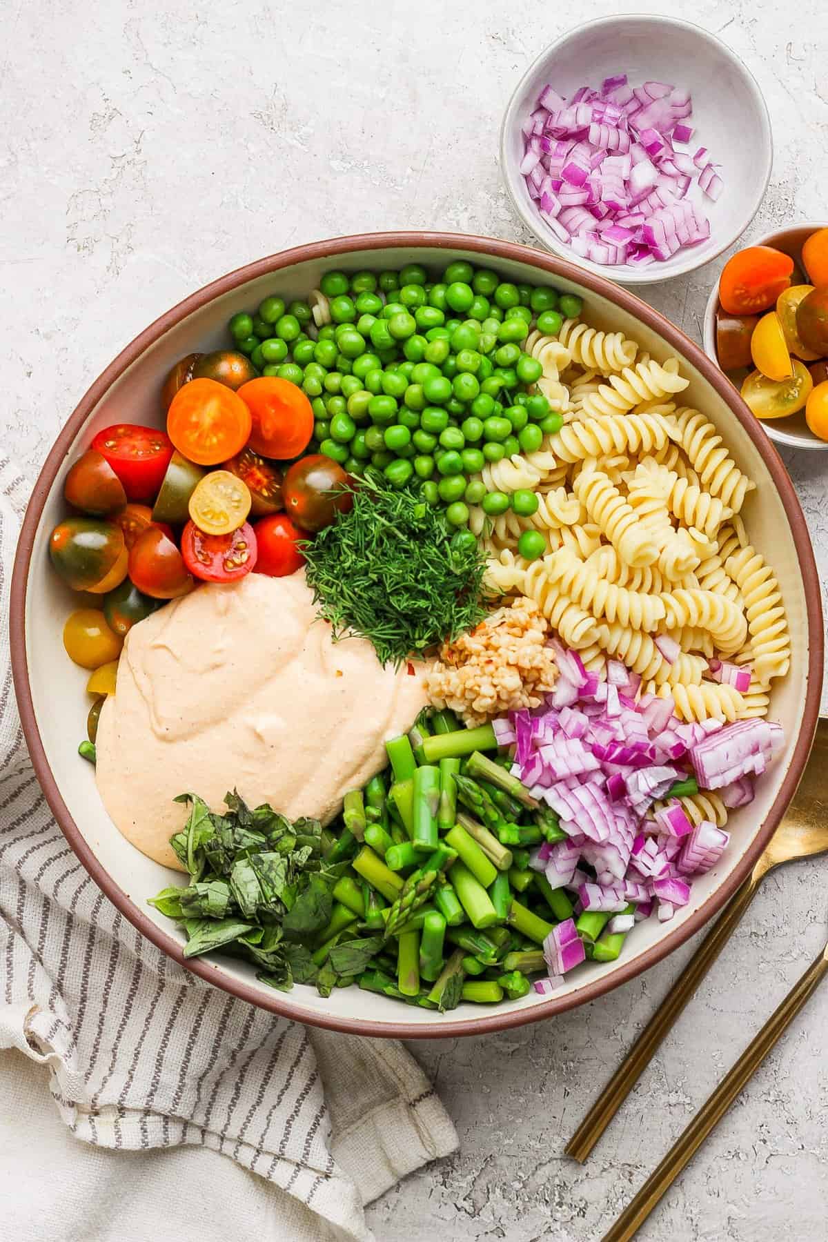 A bowl with rotini pasta, cherry tomatoes, peas, chopped red onion, green beans, fresh herbs, and a creamy sauce. A small bowl of onions and cutlery are beside it.