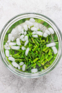 Chopped asparagus in a glass bowl of ice water, with ice cubes floating on top, against a textured white background.