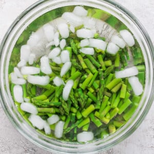 Chopped asparagus in a glass bowl of ice water, with ice cubes floating on top, against a textured white background.