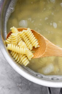 A wooden spoon holding cooked rotini pasta above a pot of boiling water.