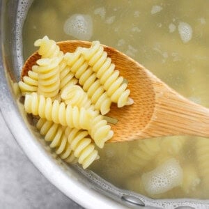 A wooden spoon holding cooked rotini pasta above a pot of boiling water.