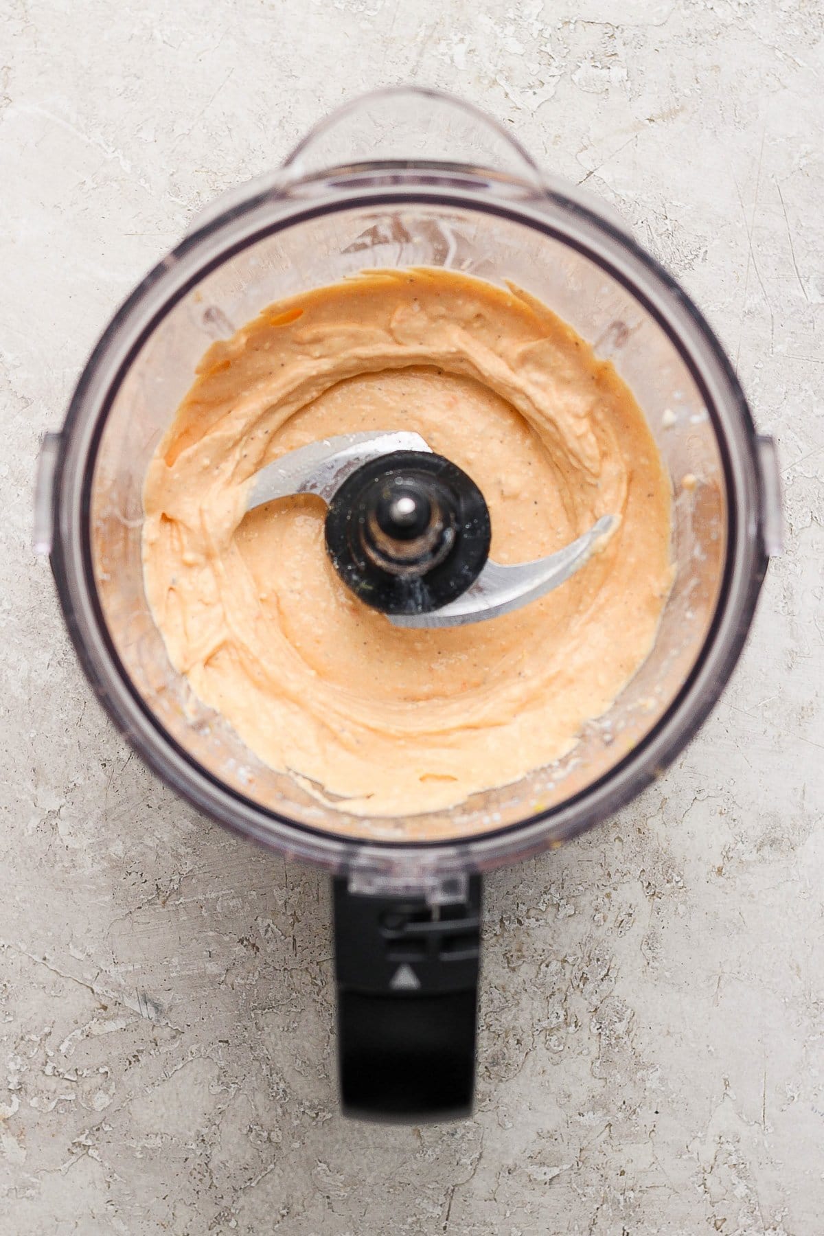 Food processor with creamy orange hummus on a gray textured surface.