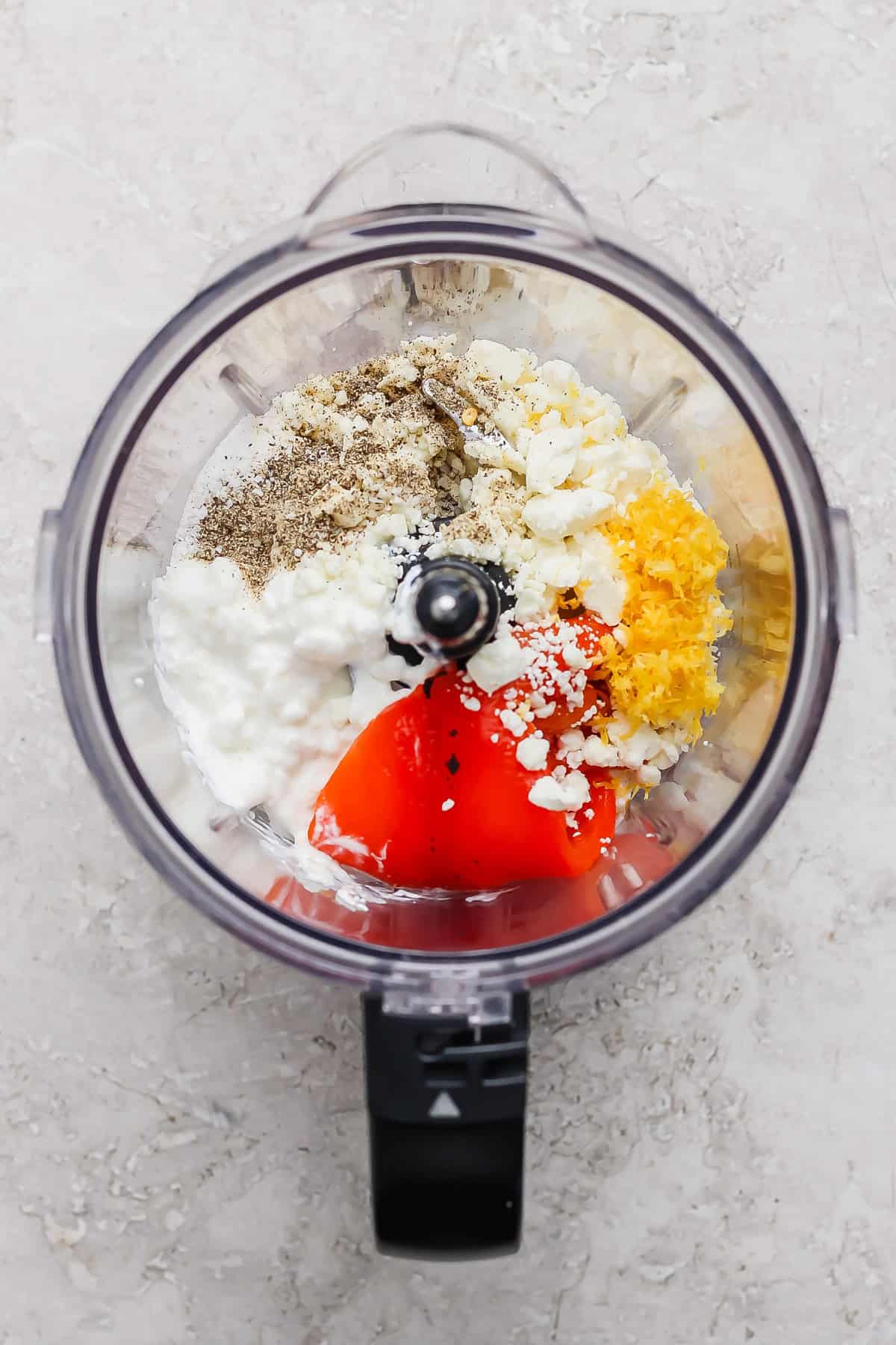Ingredients including pepper, feta cheese, cottage cheese, lemon zest, and red peppers in a food processor, ready to be blended.