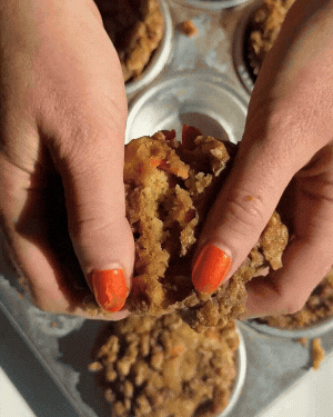 Hands with orange nail polish gently split an apple crumble muffin over a tray filled with cottage cheese carrot cake muffins.