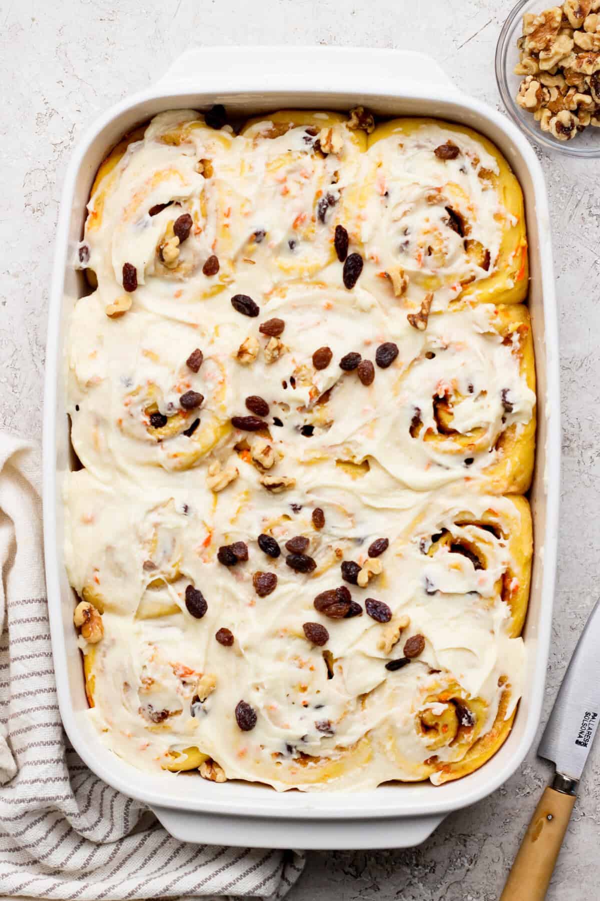 A baking dish filled with frosted carrot cake cinnamon rolls topped with raisins and walnuts. A knife and striped cloth are nearby.