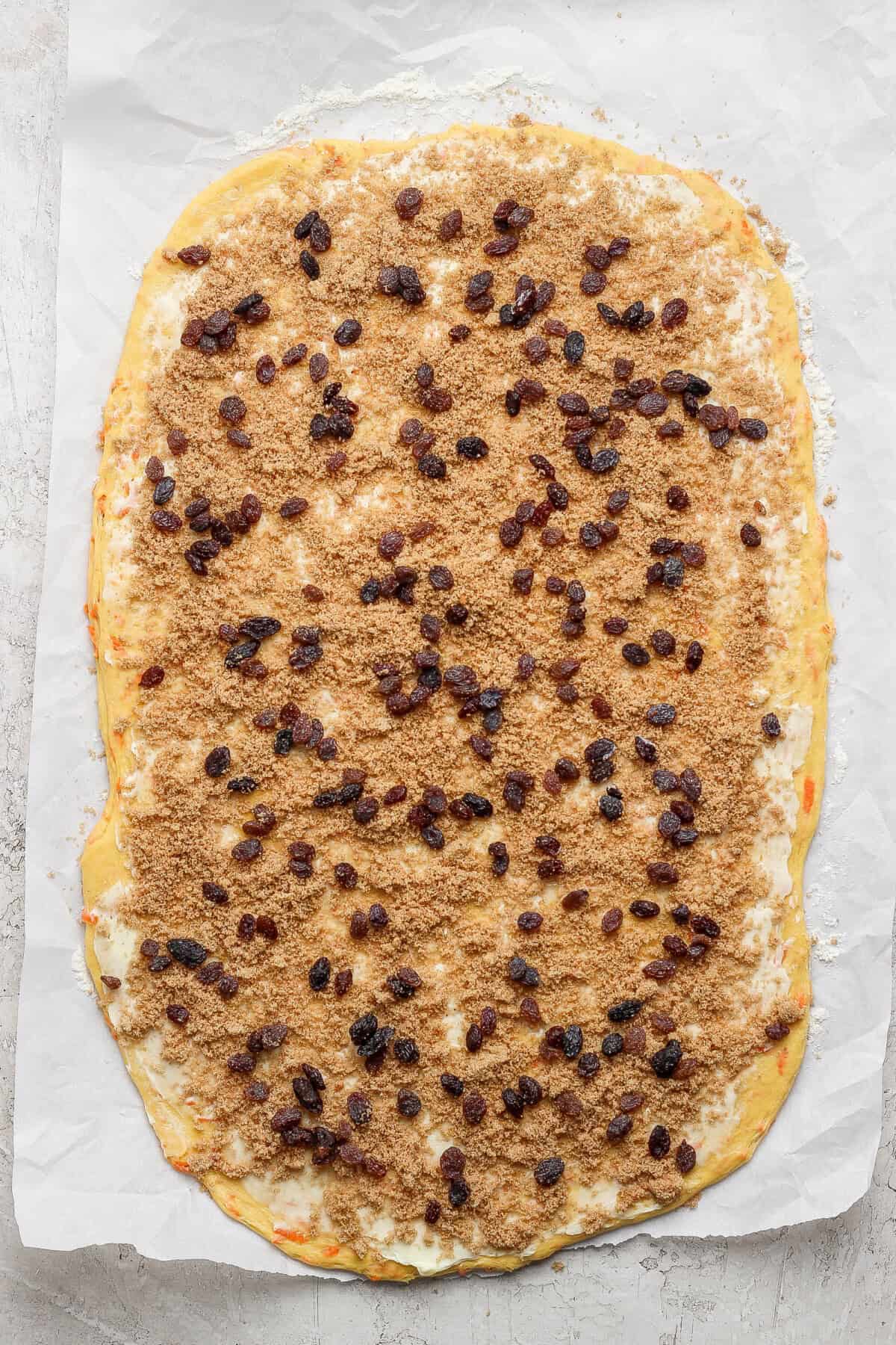 The rectangular dough on parchment paper, kissed with brown sugar and raisins, stands ready to be rolled into delightful carrot cake cinnamon rolls.