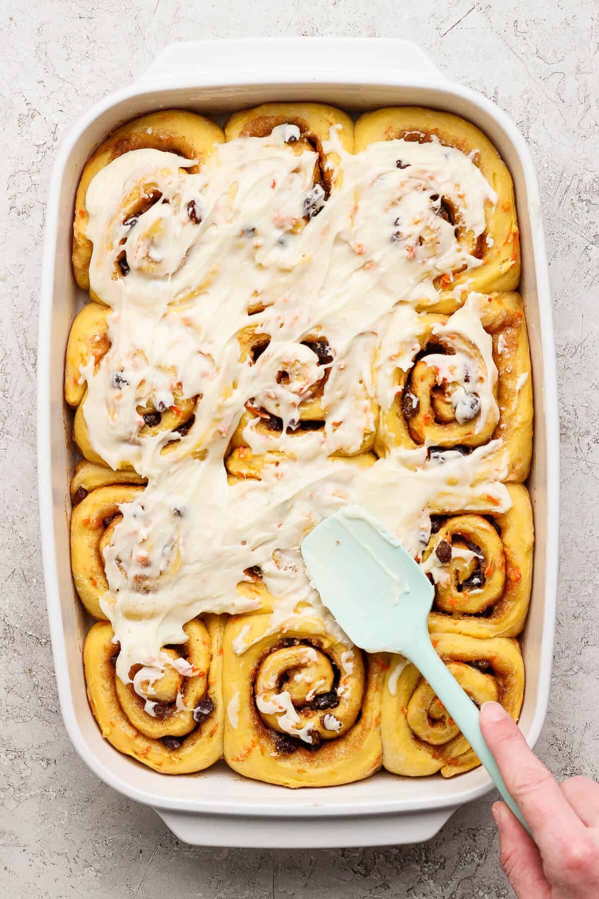 A person spreads white icing over freshly baked carrot cake cinnamon rolls arranged in a rectangular baking dish.