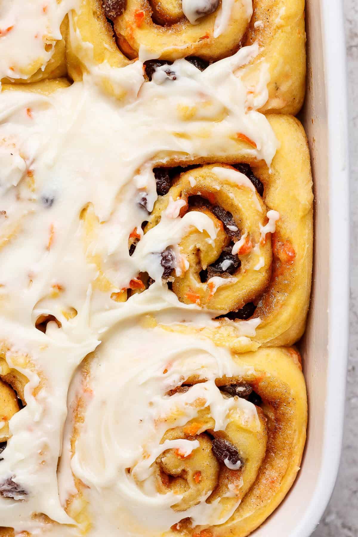 Close-up of frosted carrot cake cinnamon rolls in a baking dish, featuring swirls of dough with cinnamon and raisin filling, topped with luscious cream cheese icing.