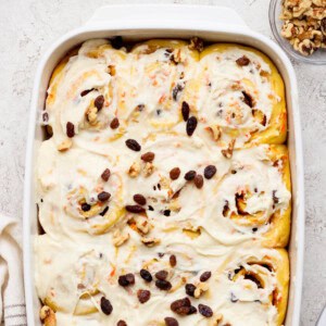 A baking dish filled with frosted carrot cake cinnamon rolls, topped with raisins and walnuts. A small bowl of walnuts is nearby.