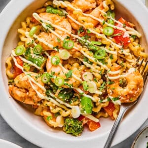 A bowl of pasta with shrimp, broccoli, red bell peppers, and green onions, drizzled with a creamy sauce and served with a fork.