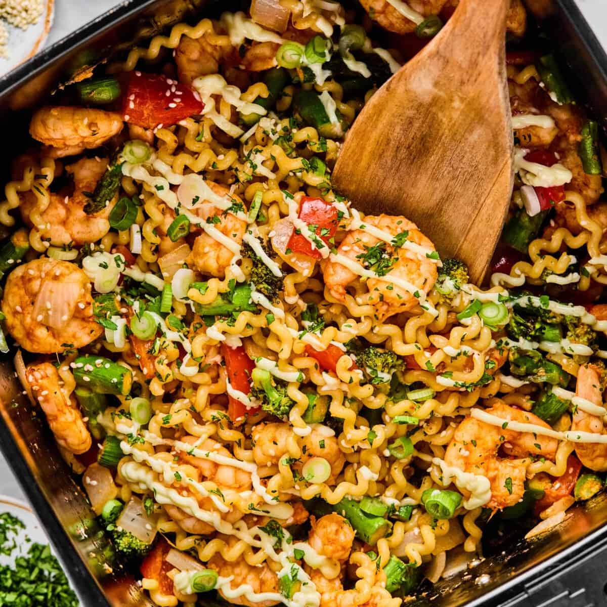 A pan filled with cooked pasta, shrimp, vegetables, and herbs being stirred with a wooden spatula.
