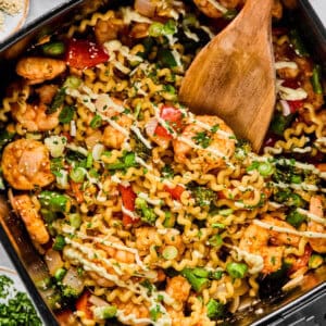 A pan filled with cooked pasta, shrimp, vegetables, and herbs being stirred with a wooden spatula.