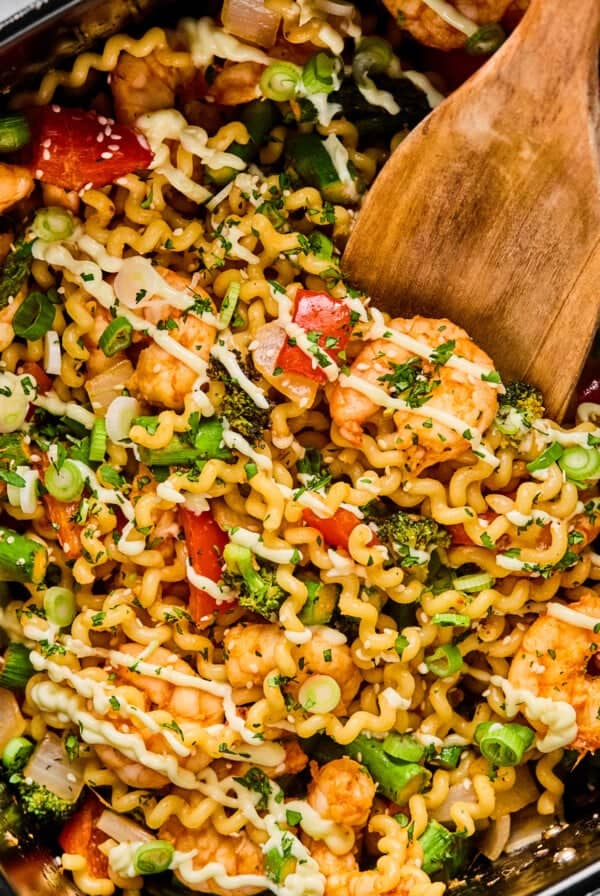 A pan filled with cooked pasta, shrimp, vegetables, and herbs being stirred with a wooden spatula.