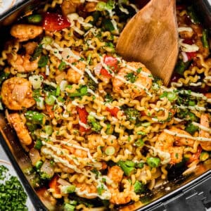 A wooden spoon stirs a mix of pasta, shrimp, vegetables, and herbs in an air fryer basket, with bowls of chopped green onions and herbs nearby.
