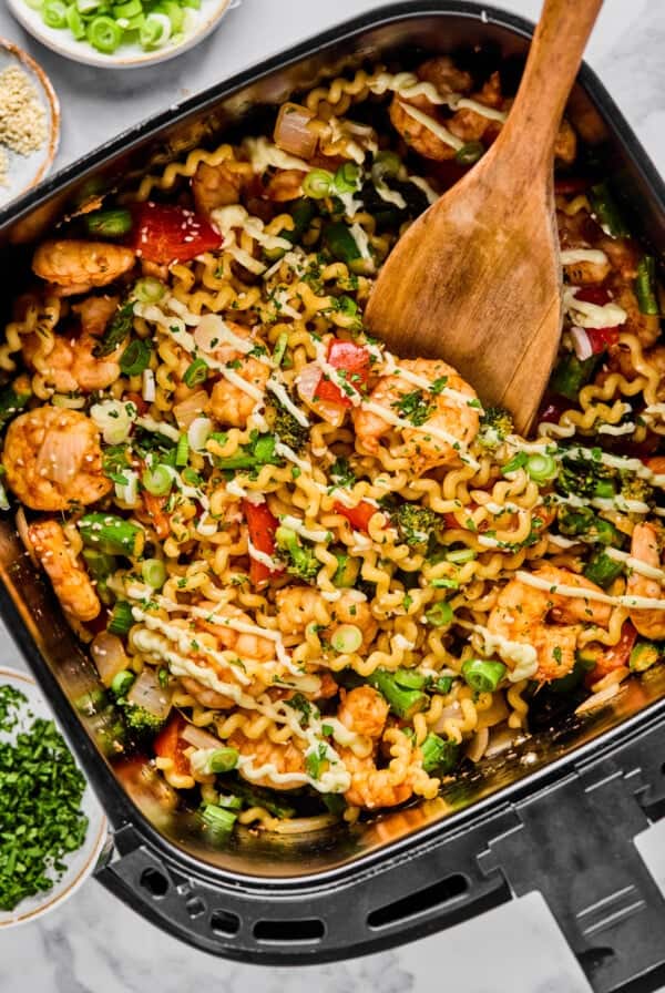 A wooden spoon stirs a mix of pasta, shrimp, vegetables, and herbs in an air fryer basket, with bowls of chopped green onions and herbs nearby.