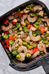 Raw shrimp, chopped broccoli, asparagus, red bell peppers, and onions arranged in an air fryer basket, ready to be cooked.