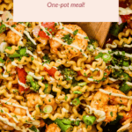 A close-up of an air fryer filled with cooked pasta, shrimp, broccoli, tomatoes, and green onions, garnished with a light drizzle of sauce. A wooden spoon rests in the dish.