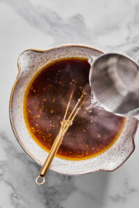 A metal whisk rests in a bowl of dark liquid while water is being poured in from a metal pitcher, all on a marble surface.