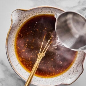 A metal whisk rests in a bowl of dark liquid while water is being poured in from a metal pitcher, all on a marble surface.