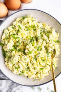 A bowl of egg salad with chopped green onions and dill, seasoned with black pepper. Three whole eggs are in the background.