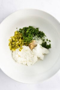 White bowl containing cottage cheese, chopped pickles, fresh dill, minced garlic, salt, and sour cream.