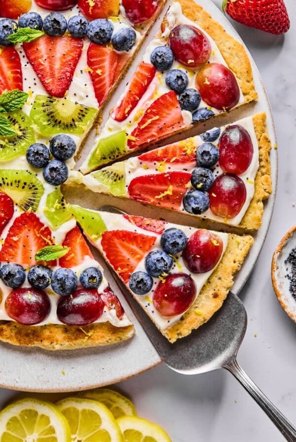 A fruit pizza topped with sliced strawberries, grapes, kiwi, and blueberries on a cream base, with a pie server lifting one slice from the plate.