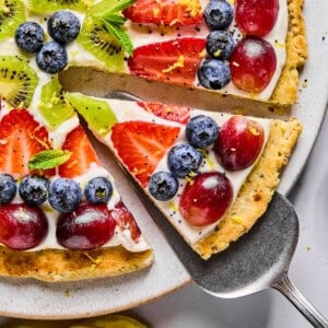 A slice of fruit pizza topped with blueberries, strawberries, grapes, kiwi, mint leaves, and lemon zest on a cookie crust with cream cheese frosting.
