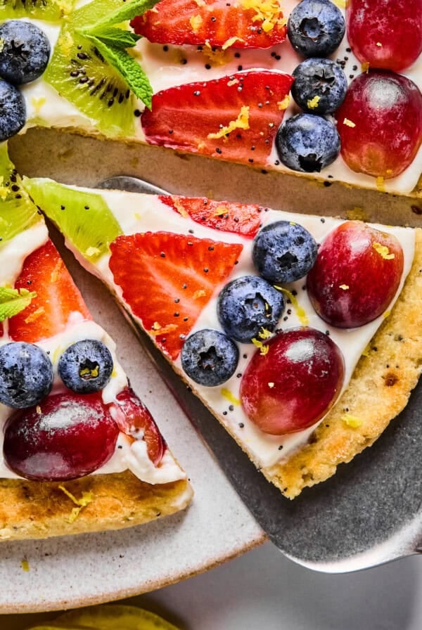 A slice of fruit pizza topped with blueberries, strawberries, grapes, kiwi, mint leaves, and lemon zest on a cookie crust with cream cheese frosting.