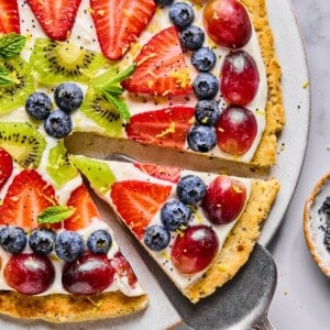 A fruit pizza topped with sliced strawberries, kiwi, blueberries, and red grapes on a creamy layer, with one slice being lifted from the whole.