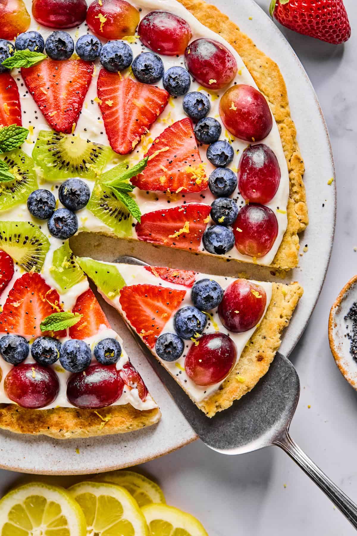 A fruit pizza topped with sliced strawberries, kiwi, blueberries, and red grapes on a creamy layer, with one slice being lifted from the whole.
