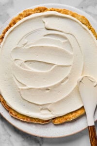 A partially frosted round cake on a plate, with a spatula spreading white frosting over the surface.