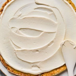 A partially frosted round cake on a plate, with a spatula spreading white frosting over the surface.