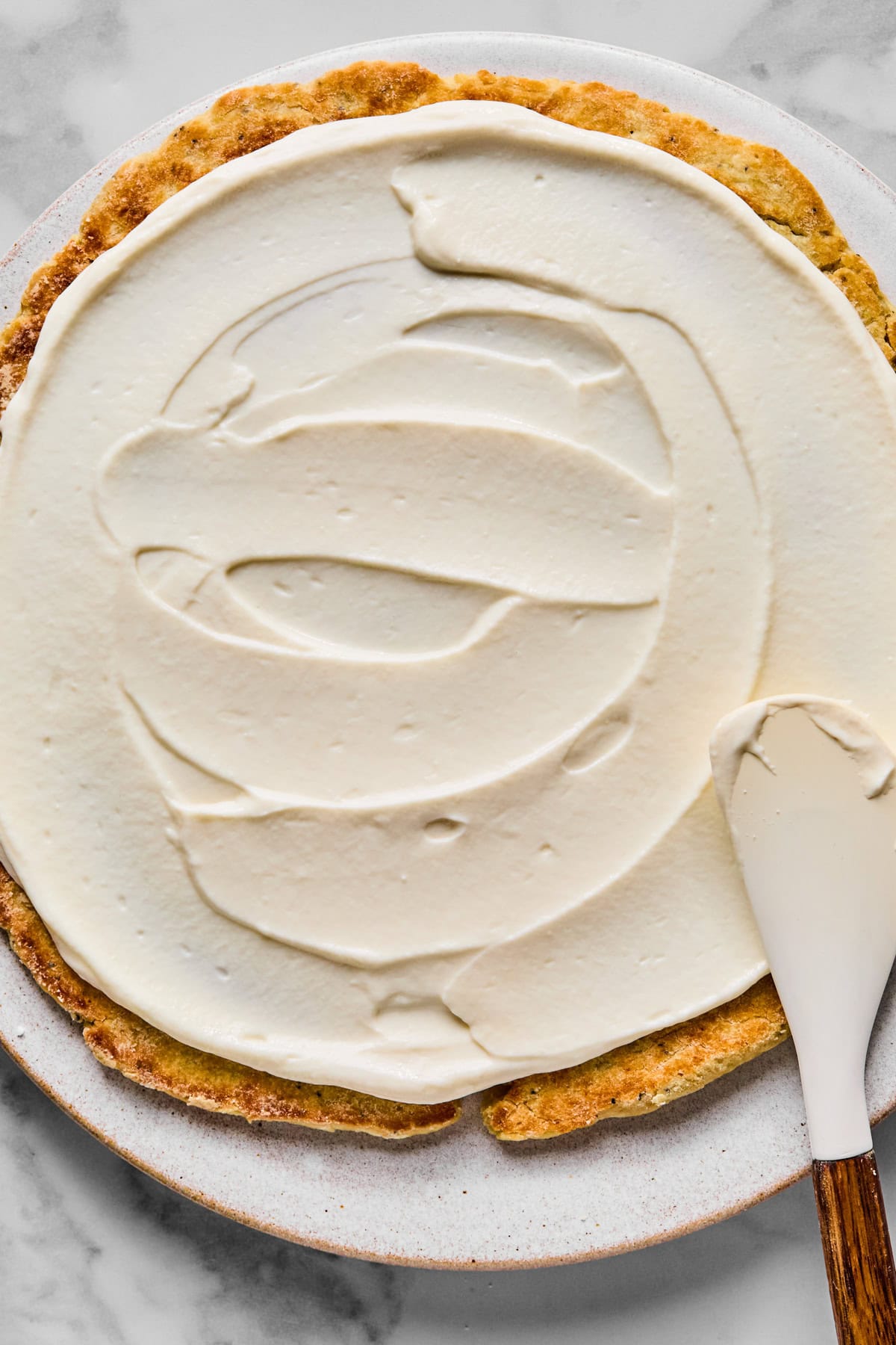 A partially frosted round cake on a plate, with a spatula spreading white frosting over the surface.