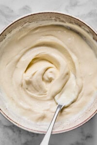 A bowl filled with smooth, creamy white yogurt being stirred with a metal spoon on a light surface.