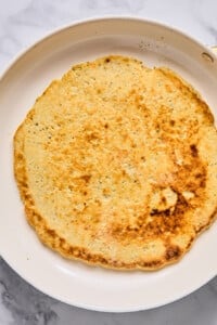 A cooked flatbread with a golden-brown surface sits in a white skillet on a marble countertop.