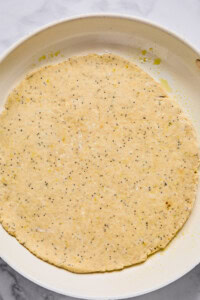 Unbaked round dough with poppy seeds spread evenly in a white baking dish on a marble surface.