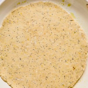 Unbaked round dough with poppy seeds spread evenly in a white baking dish on a marble surface.