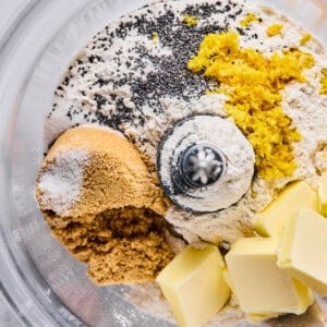 A food processor bowl containing flour, brown sugar, poppy seeds, lemon zest, salt, and cubes of butter, ready to be mixed.